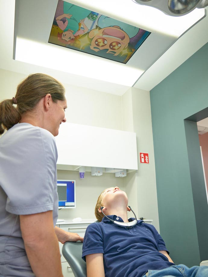 Zahnärztin behandelt ein Kind, während diese Kopfhörer trägt und eine Kinderserie auf der Decke ansieht.
